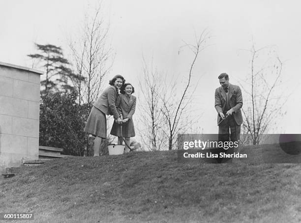 Princess Margaret Rose and Princess Elizabeth pumping water from a bucket as their father, King George VI wields the hose, in the grounds of the...