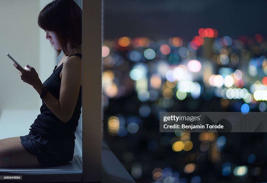 Japanese woman sitting with back to corner high-rise window using smartphone