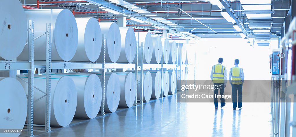 Workers in reflective clothing walking along large paper spools in printing plant