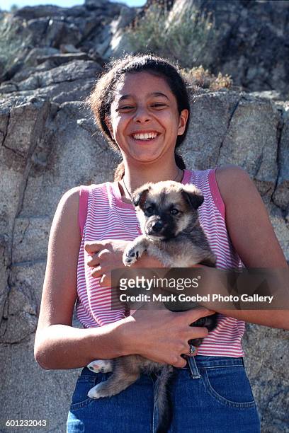 Portrait of a brown-haired teen girl as she holds a puppy and smiles, Juarez, Mexico, late 1980s. She wears blue jeans and a pink, cutoff t-shirt.