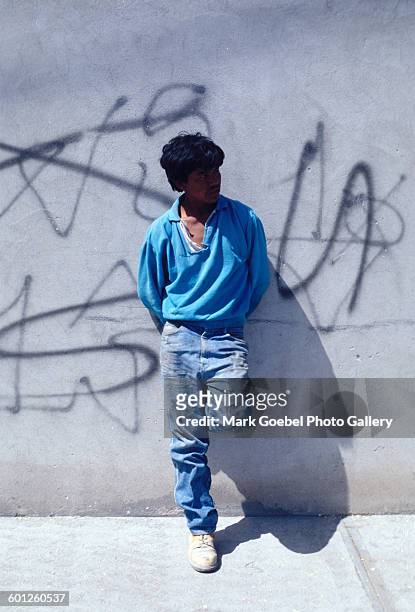 Portrait of a teen boy amputee as he stands in front of a graffiti-tagged wall, Juarez, Mexico, late 1980s. He wears a blue shirt and jeans.