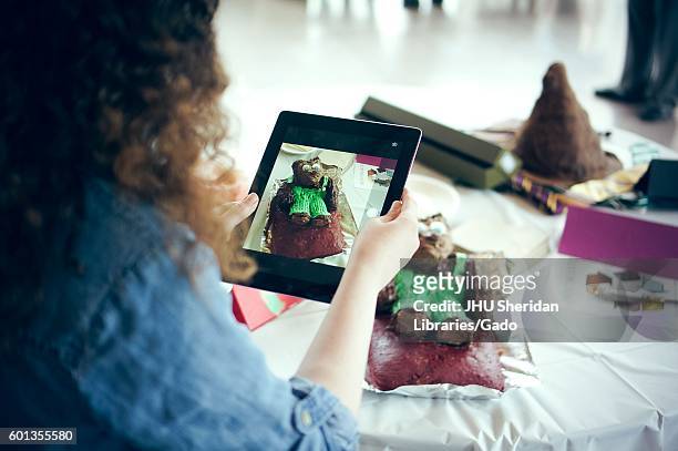 Woman taking a picture of a cake decorated for "Corduroy" by Don Freeman with an ipad at the Edible Book Festival at Johns Hopkins University,...