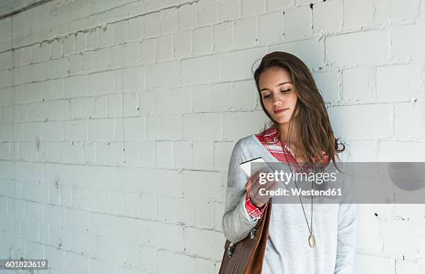 young woman leaning against brick wall looking at cell phone - looking in bag stock-fotos und bilder