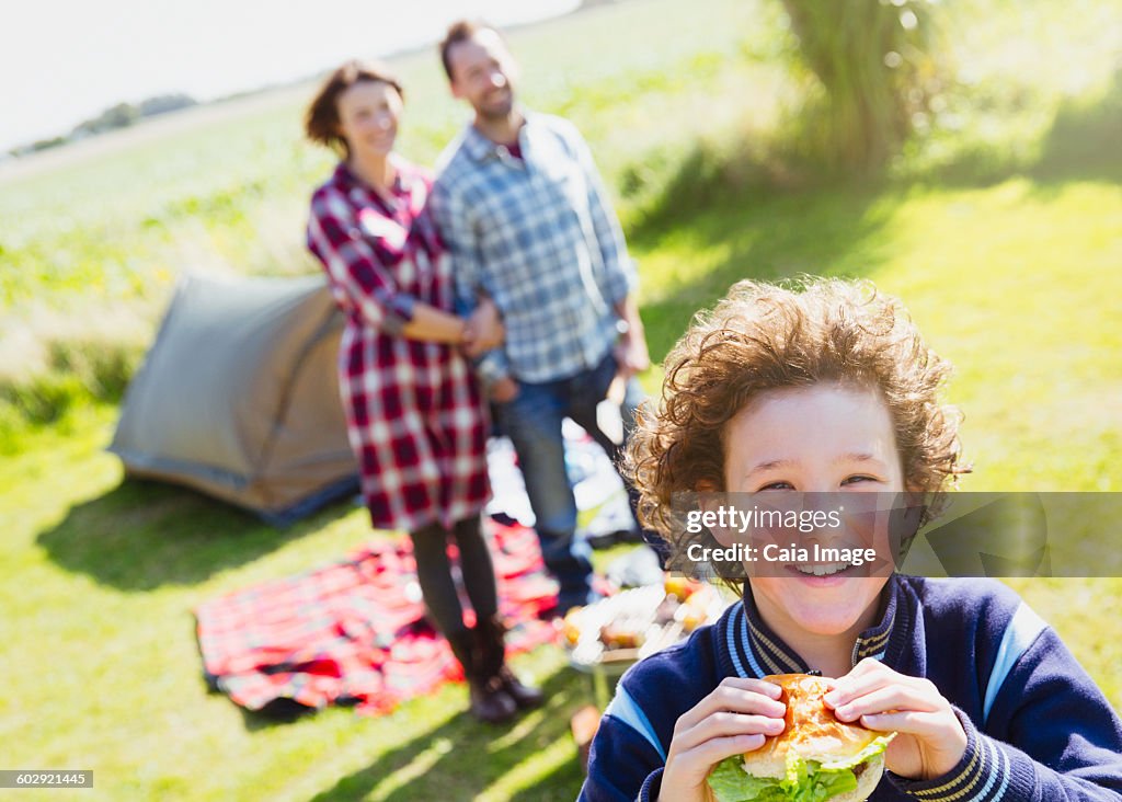 Portrait smiling boy eating hamburger with parents at sunny campsite