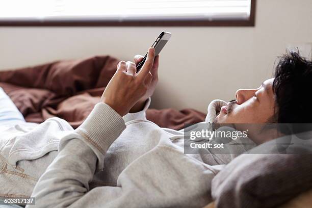 young man using smart phone lying on bed - sweatshirt stock-fotos und bilder