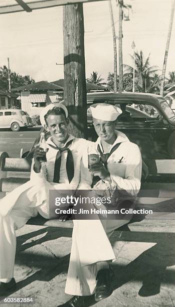 Two sailors sitting on bench drinking beer or soda from bottles. In background are parked cars and palm trees in residential area.