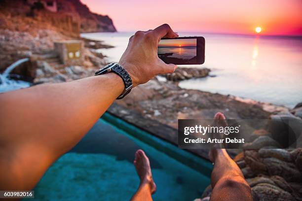 guy from personal perspective taking pictures with smartphone from a natural pool in the mediterranean sea costa brava shoreline watching the sunrise with his legs levitating in the air during summer time. - amazing moment in the nature stock-fotos und bilder