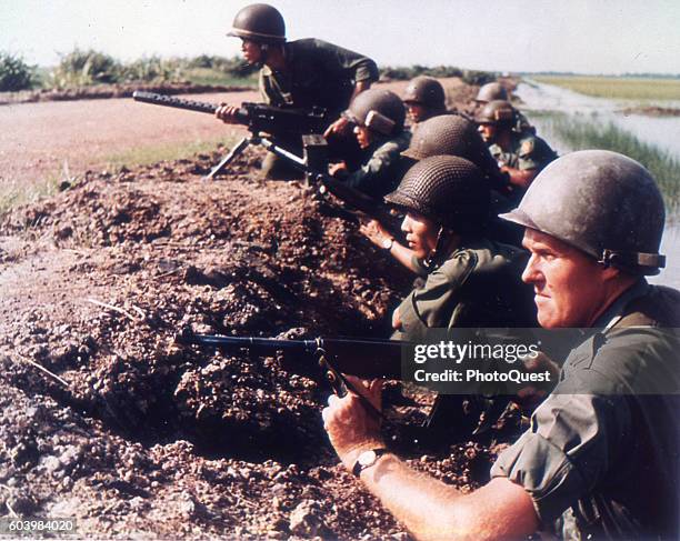 Along the bank of a rice paddy, a US Army advisor and a line of Vietnamese infantrymen look into the distance, Vietnam, 1965.