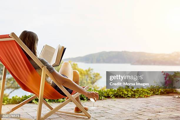 woman reading book while relaxing on deck chair - wetterfester stuhl stock-fotos und bilder