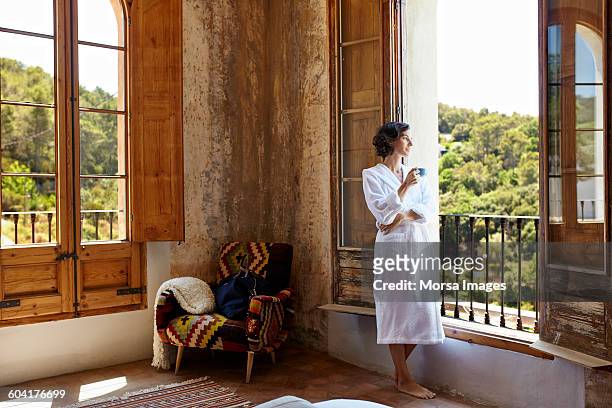 woman having coffee while standing at window - travel fotografías e imágenes de stock