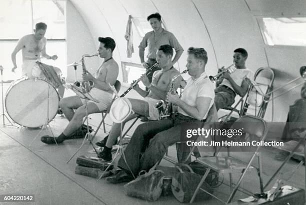 Men sitting on folding chairs playing musical instruments in quonset hut.