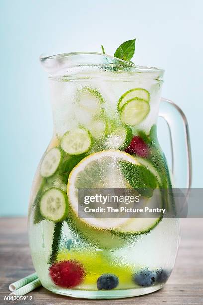 infused water with cucumber, lemon, lime, berry and mint on blue background - geldpresse stock-fotos und bilder