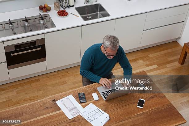 mature man in kitchen with construction plan using laptop - calculator top view stock pictures, royalty-free photos & images