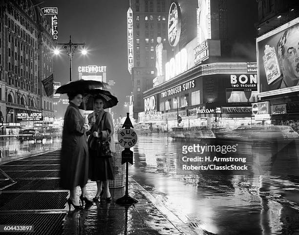 1950s TWO SILHOUETTED WOMEN AT BUS STOP UNDER UMBRELLAS RAINY NIGHT TIMES SQUARE MANHATTAN NEW YORK USA