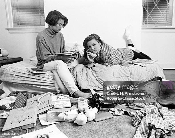 1980s TWO TEENAGE AFRICAN AMERICAN GIRLS IN CLUTTERED BEDROOM ONE TALKING ON THE PHONE ONE READING MAGAZINE