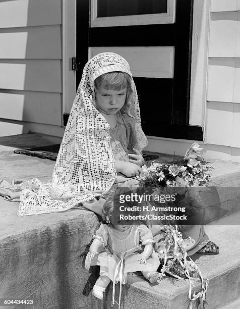 1950s SAD LITTLE GIRL IN MOCK BRIDE COSTUME WITH BOUQUET IN LAP & BABY DOLL AT FEET WITH DEJECTED LOOK ON FACE