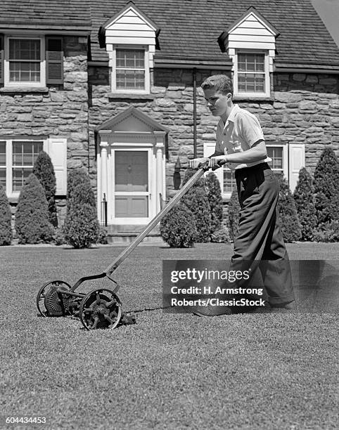 1940s TEENAGE BOY MOWING GRASS PUSHING PUSH MOWER ON FRONT LAWN OF STONE SUBURBAN HOUSE