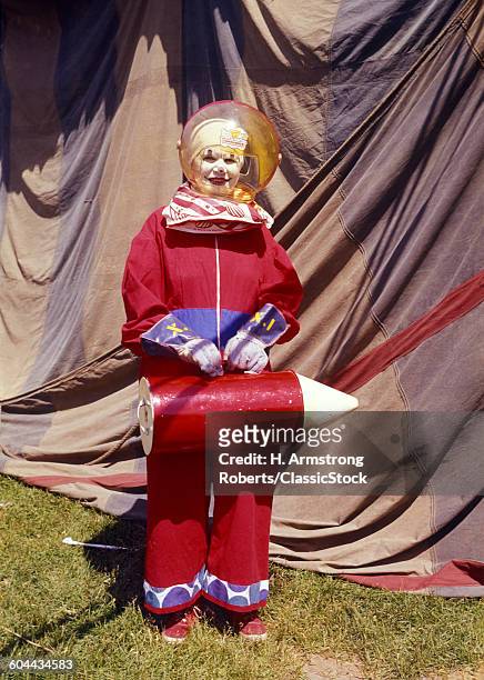 1970s CIRCUS CLOWN WEARING SPACE HELMET AND SUIT CARRYING ROCKET STANDING BY TENT LOOKING AT CAMERA