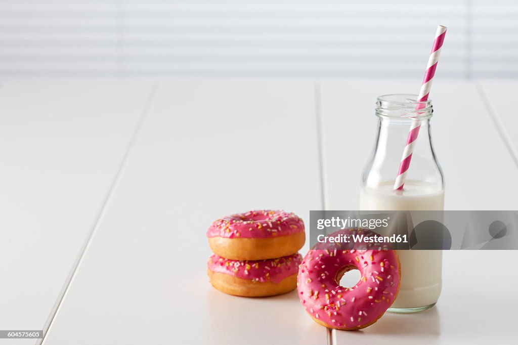Glass bottle of milk and three doughnuts with pink icing on white ground