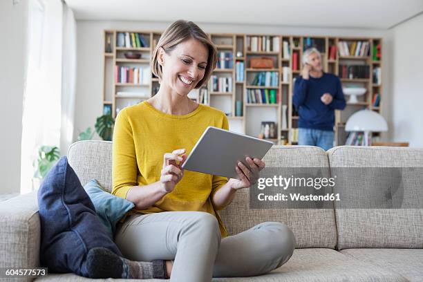 smiling woman sitting on the couch using digital tablet while her husband telephoning in the background - man middelbare leeftijd woonkamer stockfoto's en -beelden