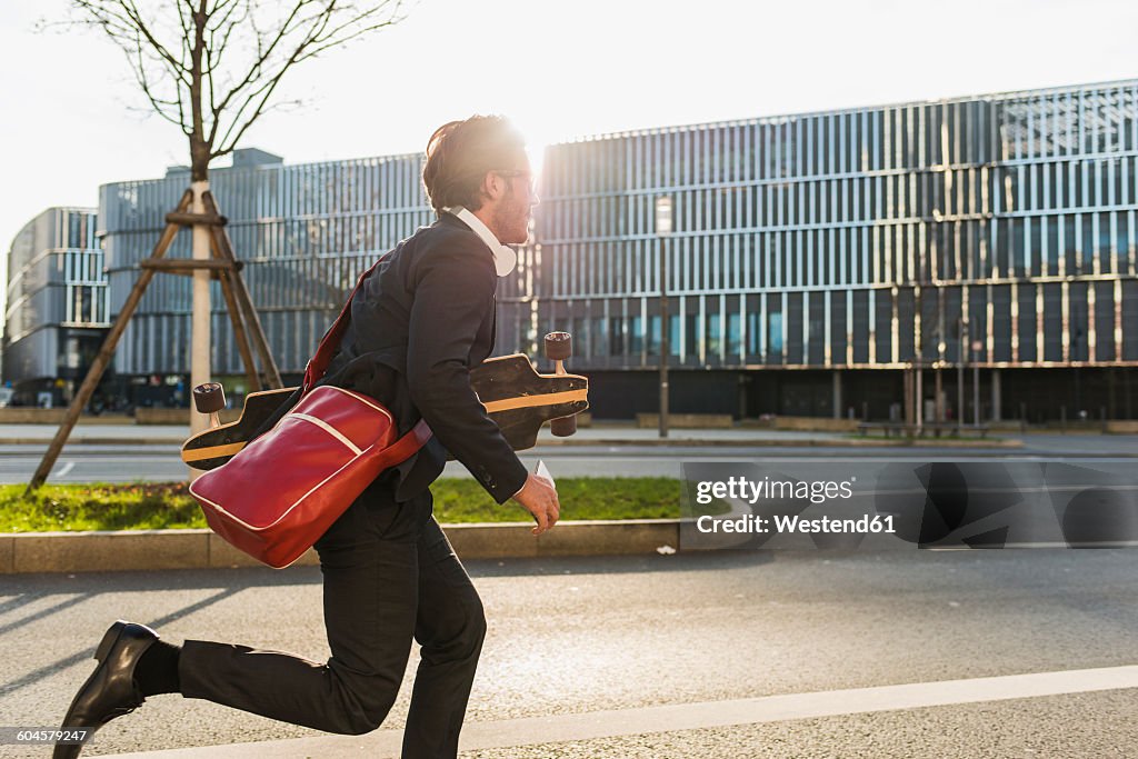 Germany, Frankfurt, Young businessman running with skateboard under his arm, using mobile phone