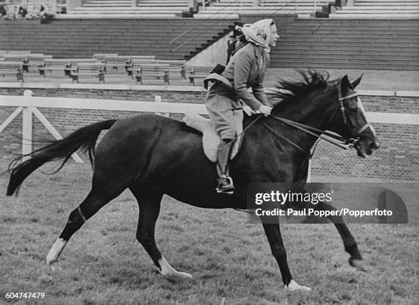 Queen Elizabeth II wears a fawn coloured jacket, breeches and head scarf as she gallops on a black horse along the course at Ascot Racecourse in...