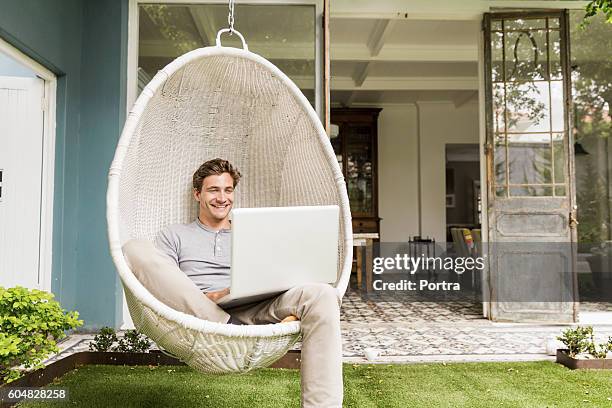 happy young man using laptop on swing chair - garden working stockfoto's en -beelden