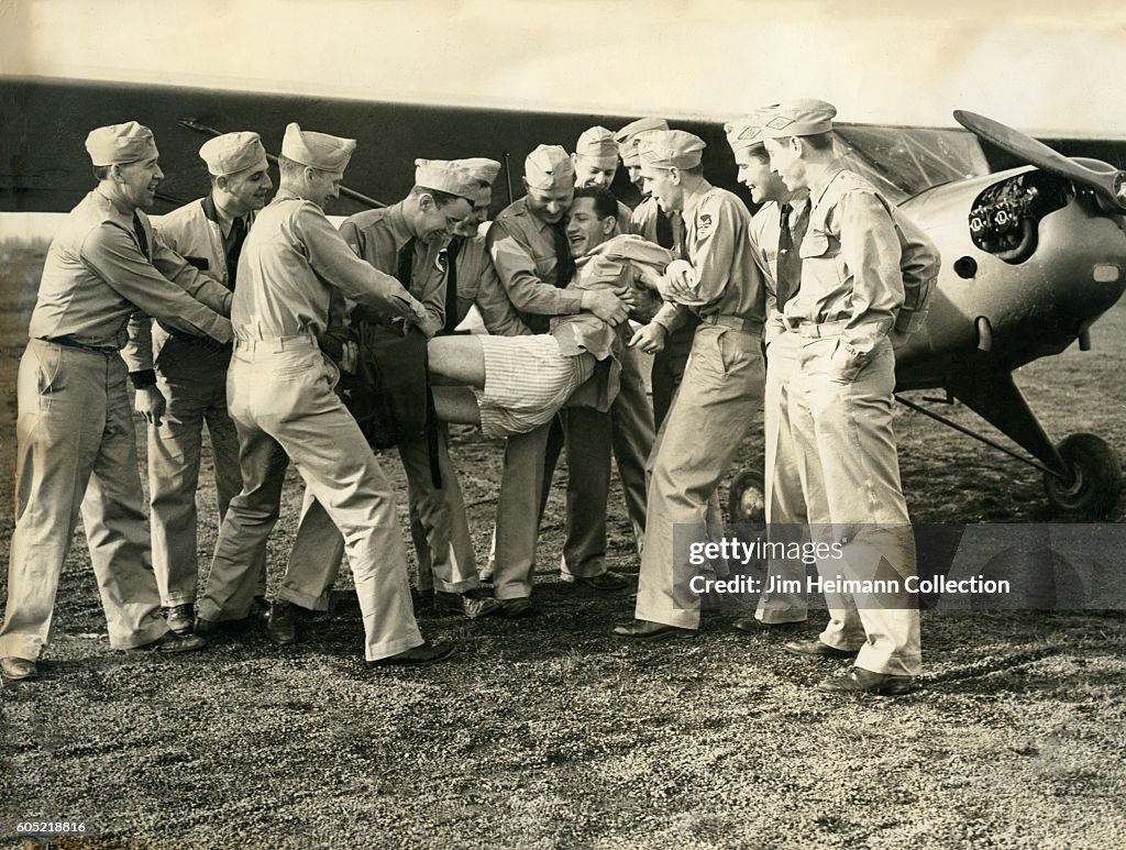 Servicemen pulling pants off man by plane.