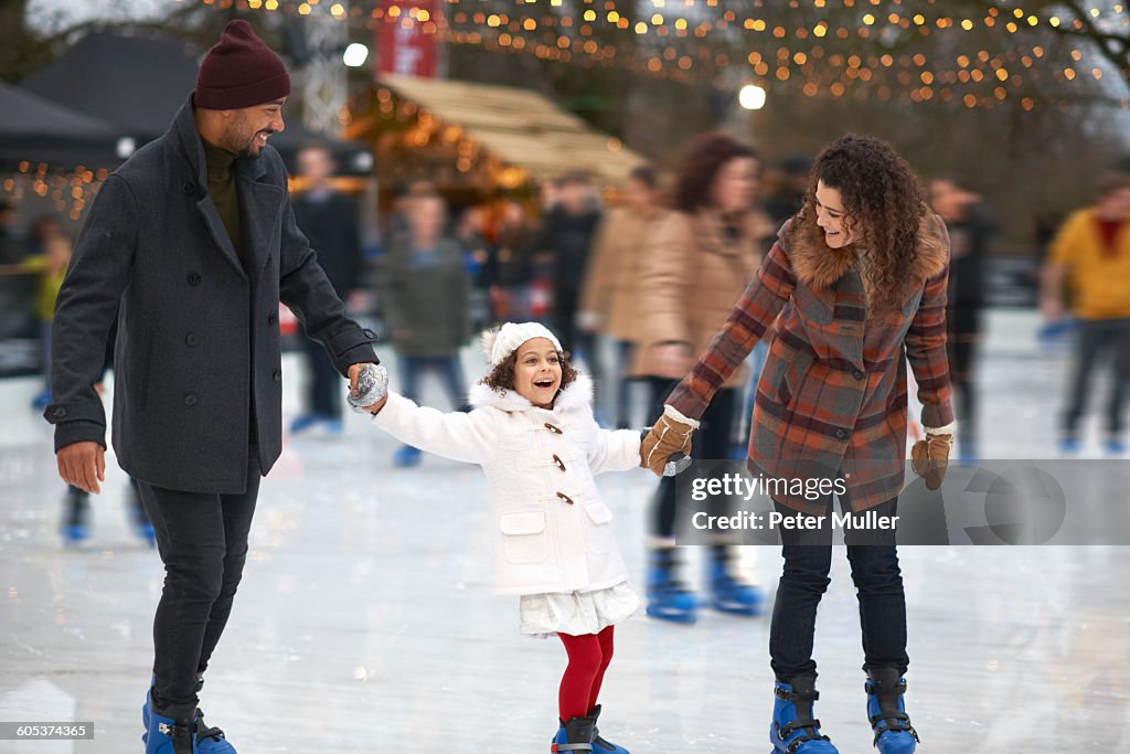 Girl holding parents hands ice skating, smiling