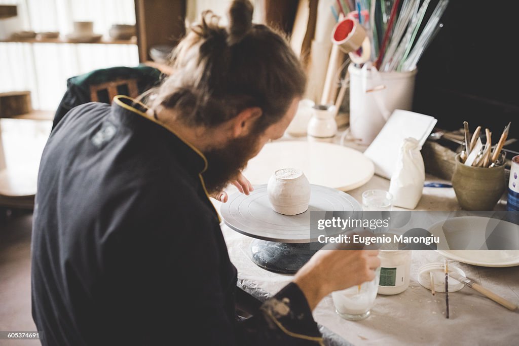 Rear view of mid adult man in workshop applying ceramic glaze to clay pot