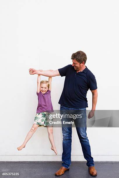 portrait of mature man with daughter hanging from his arm in front of white wall - hanging clothes stock-fotos und bilder