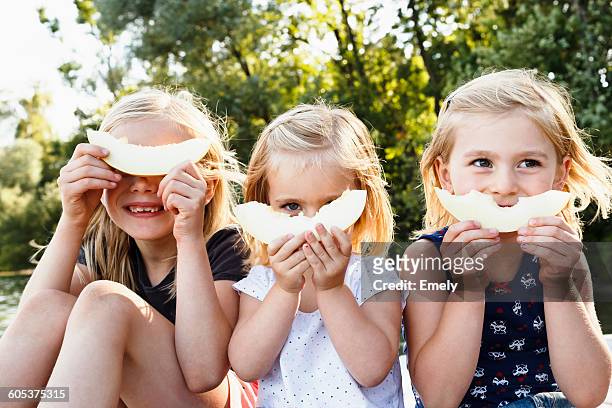 portrait of three young sisters holding smiling melon in front of face in park - children only stock-fotos und bilder