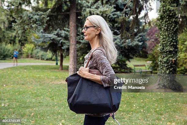 mature woman walking through park carrying shoulder bag - shoulder bag foto e immagini stock