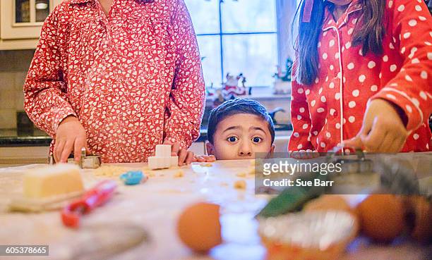boy in kitchen making cookies with sisters peeking over kitchen counter looking at camera - great american group stock pictures, royalty-free photos & images