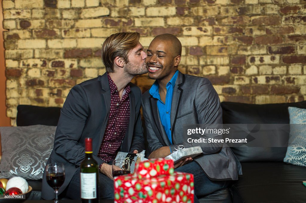 Male couple sitting on sofa, exchanging Christmas gifts, young man kissing partner on cheek
