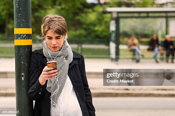 young woman listening to music through headphones using mobile phone at tram station - winter scarf stock pictures, royalty-free photos & images