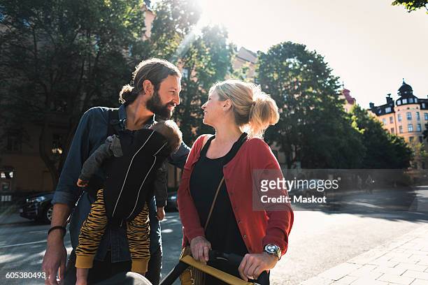 loving mid adult parents with baby boy walking on sidewalk in city - portabebés fotografías e imágenes de stock
