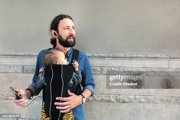 mid adult father listening music while carrying baby in carrier against wall - family looking at smartphone stockfoto's en -beelden