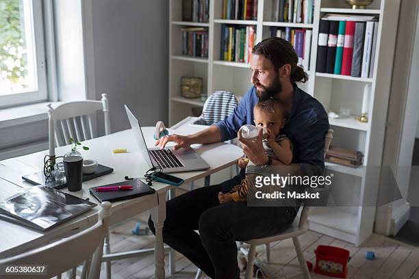 father feeding baby boy while using laptop at home - toddler milk stock-fotos und bilder