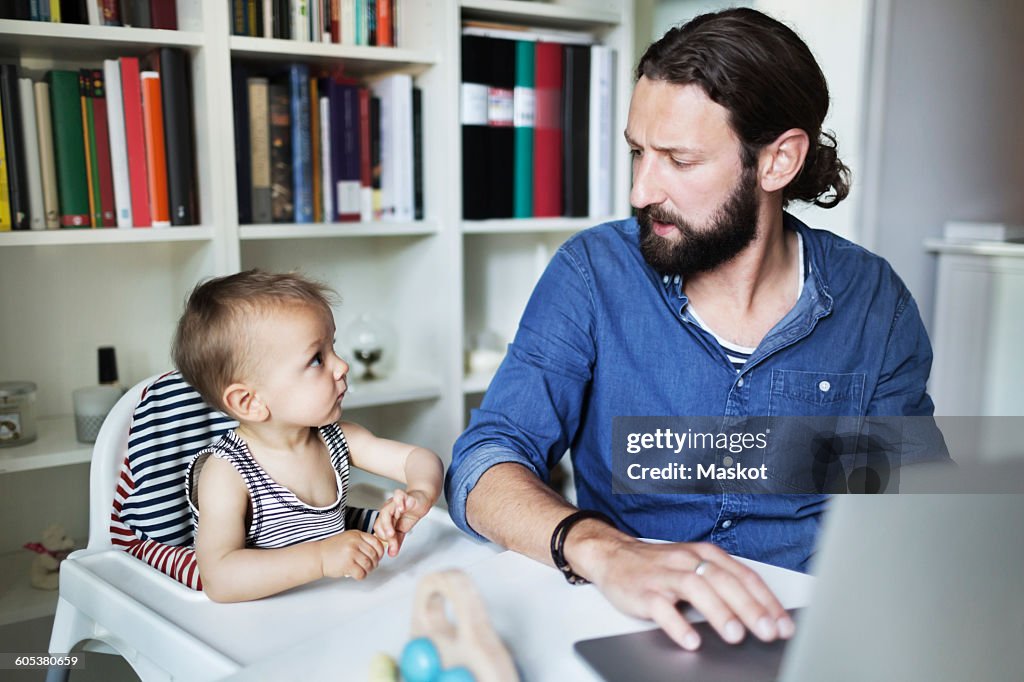 Mid adult businessman using laptop while looking at baby boy at home