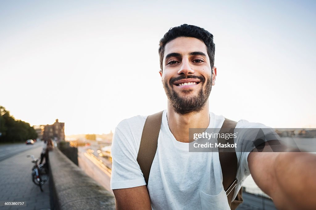 Portrait of happy male tourist sitting on retaining wall of bridge