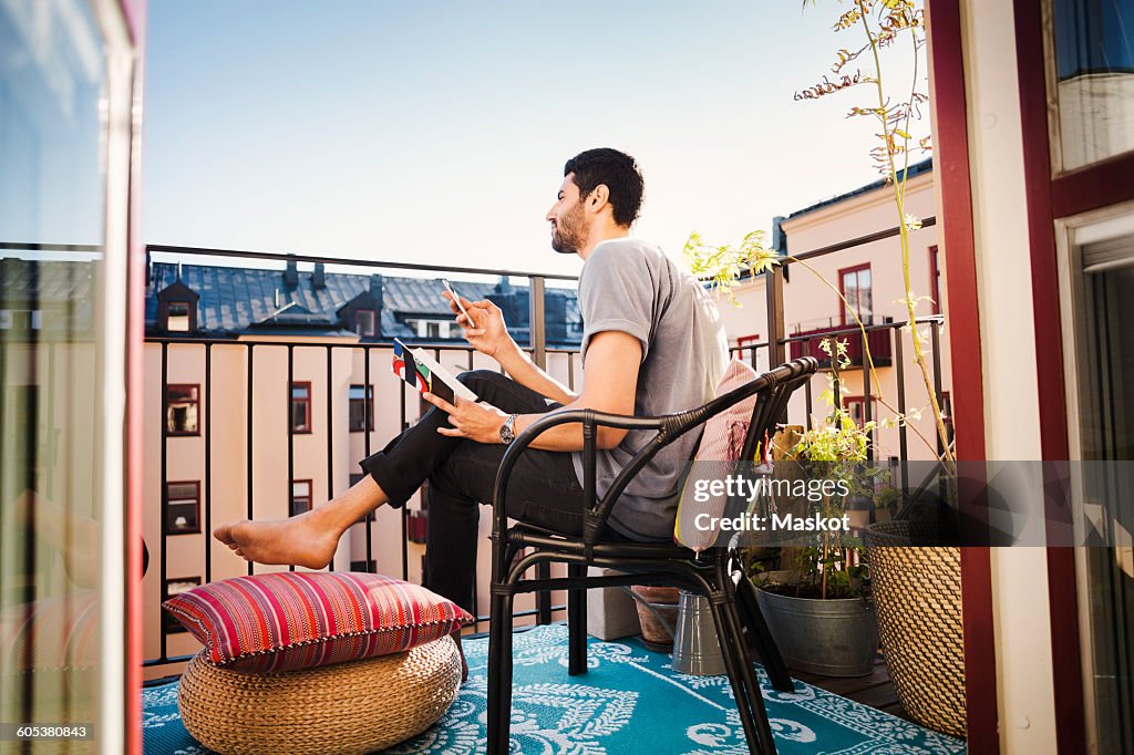 Side view of man holding smart phone and guidebook while looking away at balcony