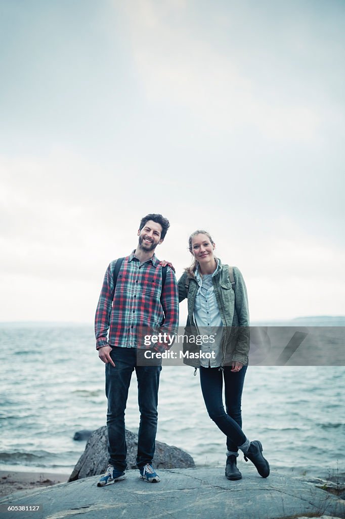 Full length portrait of confident wonderlust couple standing on rock by sea