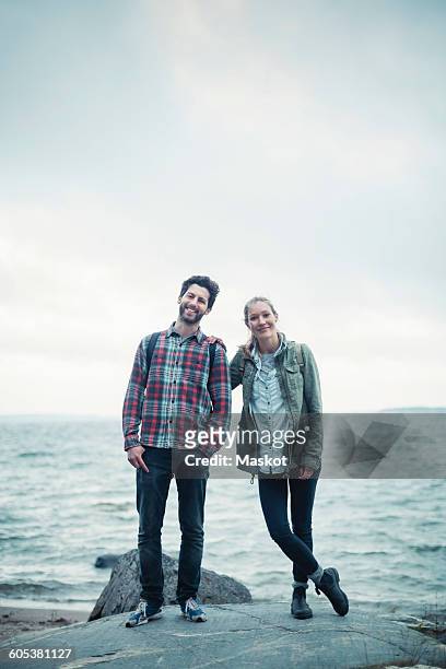 full length portrait of confident wonderlust couple standing on rock by sea - man standing full length side stock-fotos und bilder