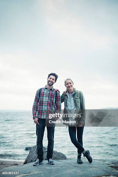 full length portrait of confident wonderlust couple standing on rock by sea - outdoor guy sitting on a rock stockfoto's en -beelden