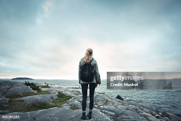 rear view of woman standing on rock by sea against cloudy sky - rücken stock-fotos und bilder