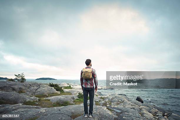 full length rear view of man standing on rock by sea against cloudy sky - man standing full length side stock-fotos und bilder