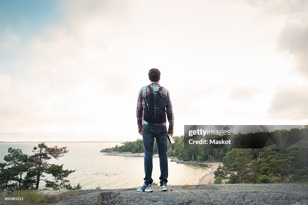 Full length of man standing on rock at lakeshore against cloudy sky