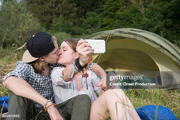 young couple kissing and taking selfie with smart phone in front of camp tent, bavaria, germany - boy taking picture in forest stock-fotos und bilder