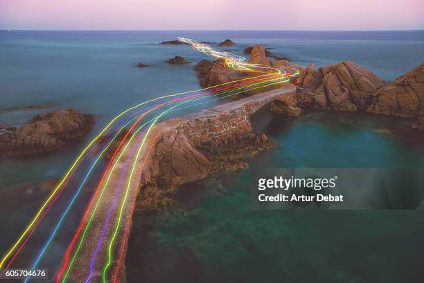 long exposure of a colorful rainbow of light traces in a beautiful curved path between the rocks over the mediterranean sea in the costa brava shoreline on sunset. - asentamiento humano fotografías e imágenes de stock
